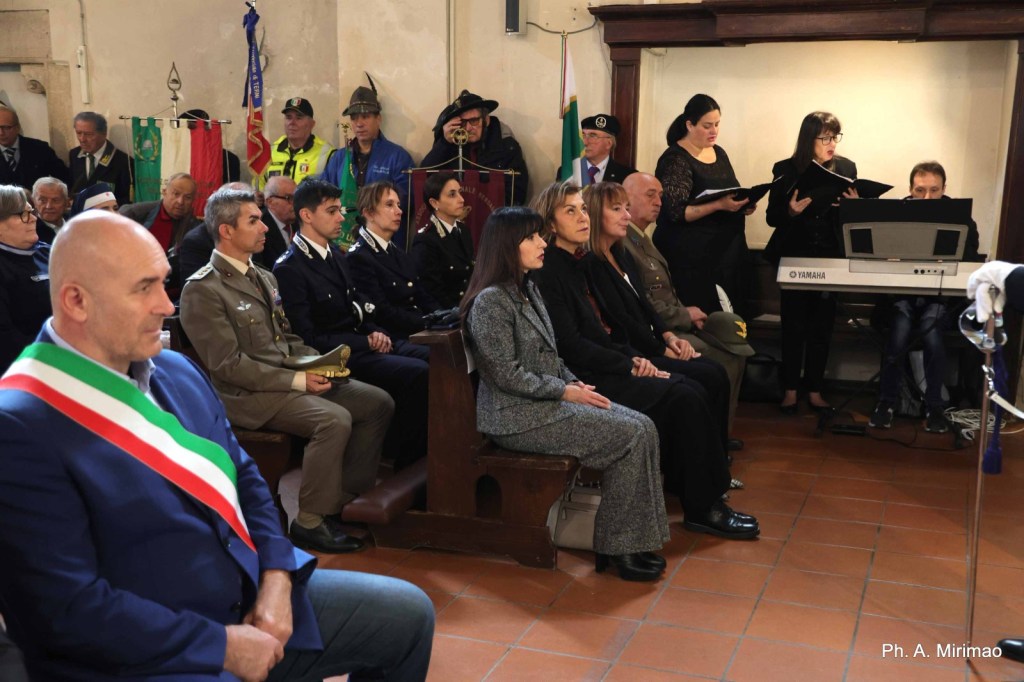 A group of people seated in a hall during a formal event, with flags and a piano in the background. Individuals in military and official attire are in attendance, while some women are singing.