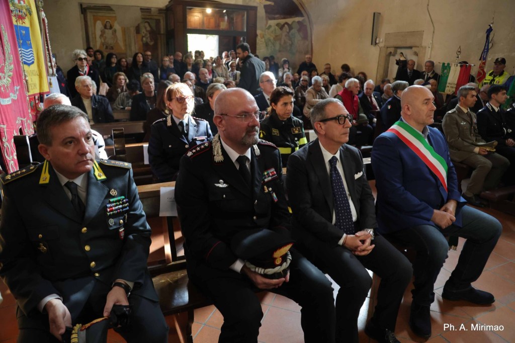 A formal event taking place indoors with a crowd gathered. Several individuals in uniform, including military and law enforcement personnel, are seated in the foreground, while more attendees can be seen in the background.