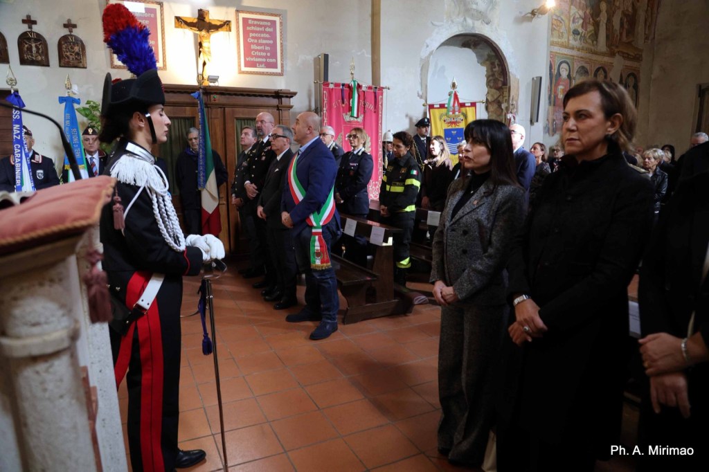 Ceremony inside a church with attendees in formal attire, including a soldier in uniform, local officials, and community members gathered for a memorial event.