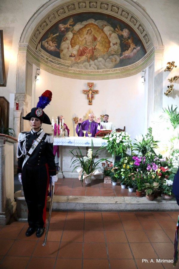 A ceremonial scene in a church with a soldier in uniform standing guard in the foreground, while a religious service takes place at the altar adorned with flowers and decorations.