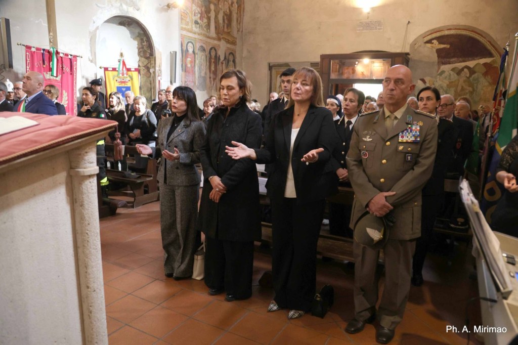 A group of people standing in a church, engaged in a solemn moment, with some members in military uniforms and others in formal attire. The interior features religious decorations and banners.