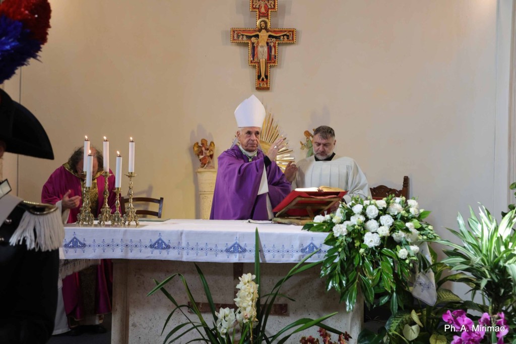 A bishop wearing a white mitre and a purple robe is performing a religious ceremony at an altar, accompanied by two other clergy members. The altar is adorned with white flowers and candles, with a crucifix and religious statues in the background.