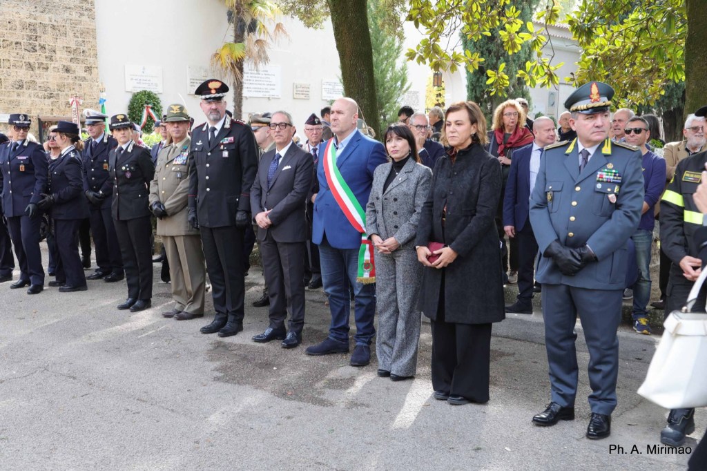 A group of formally dressed individuals stands at attention during a commemorative event. Various officials, including police and military personnel, are present, along with local dignitaries, as they pay their respects.