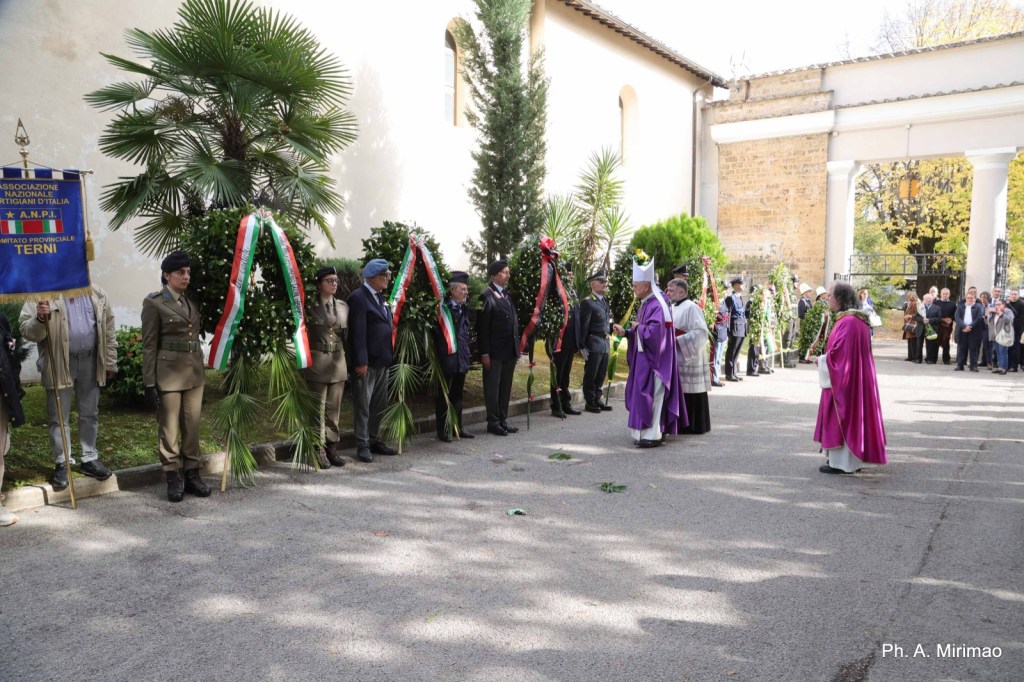 Ceremony with participants holding wreaths and banners outdoors, commemorating an event, with a priest in ceremonial attire.