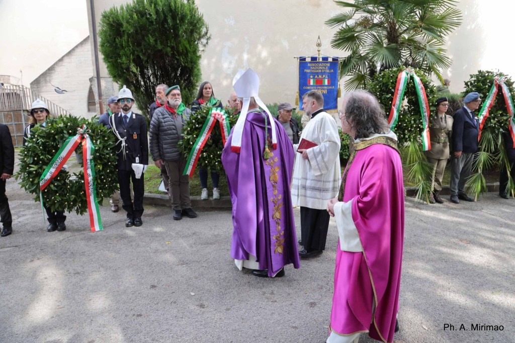 Ceremony with participants in formal attire holding wreaths, including religious figures in traditional robes, surrounded by greenery and banners.