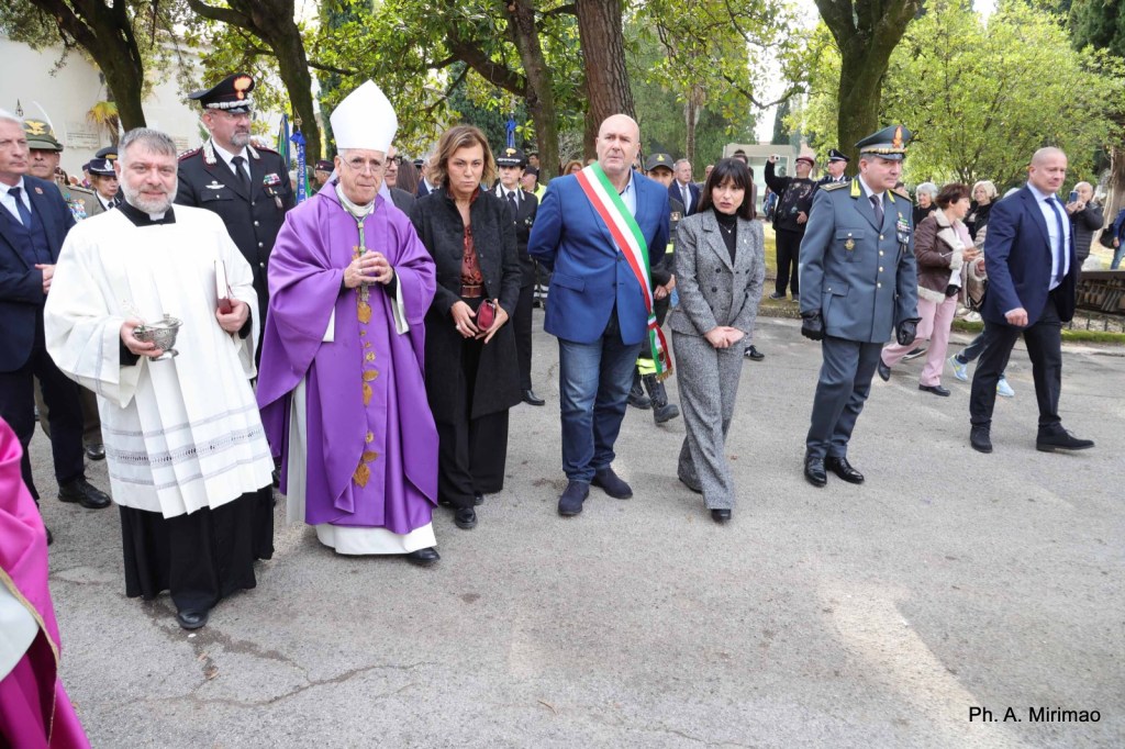 A group of officials and clergy members walking together in a park during a ceremony. The central figure is dressed in purple liturgical robes, while a local official wears a tricolor sash. Others include military personnel and civilians observing the event.
