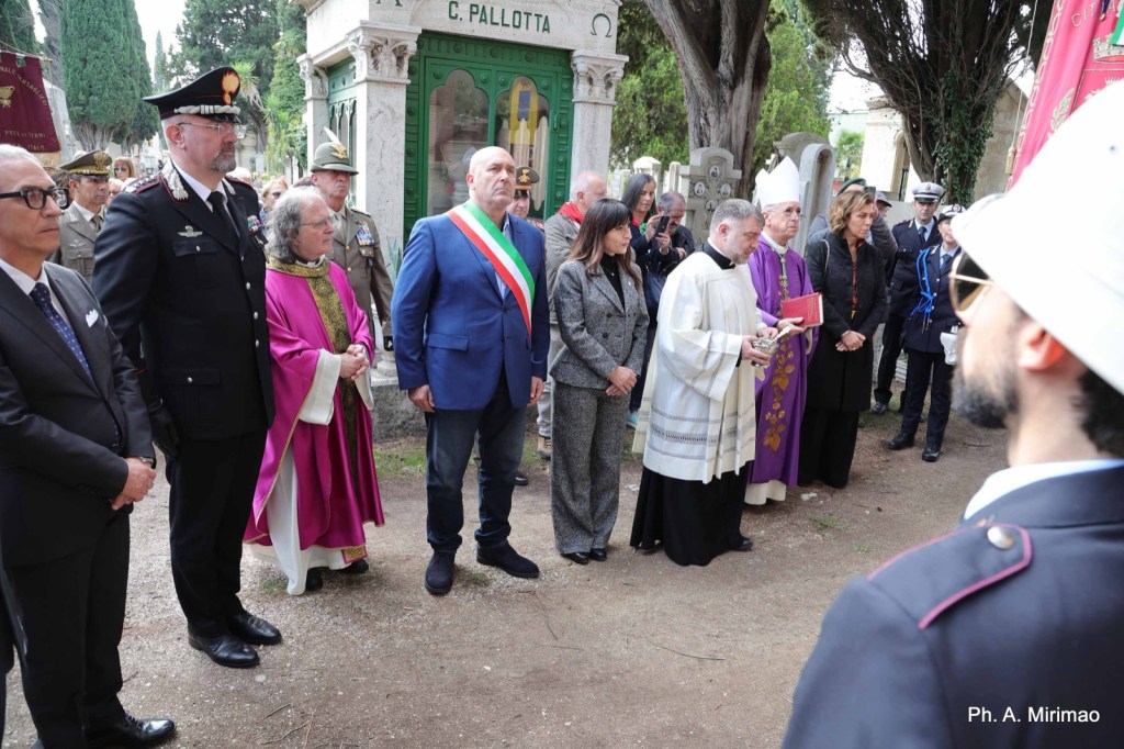 A group of people, including officials and clergy, standing solemnly during a ceremony in a cemetery, with gravestones and trees in the background.