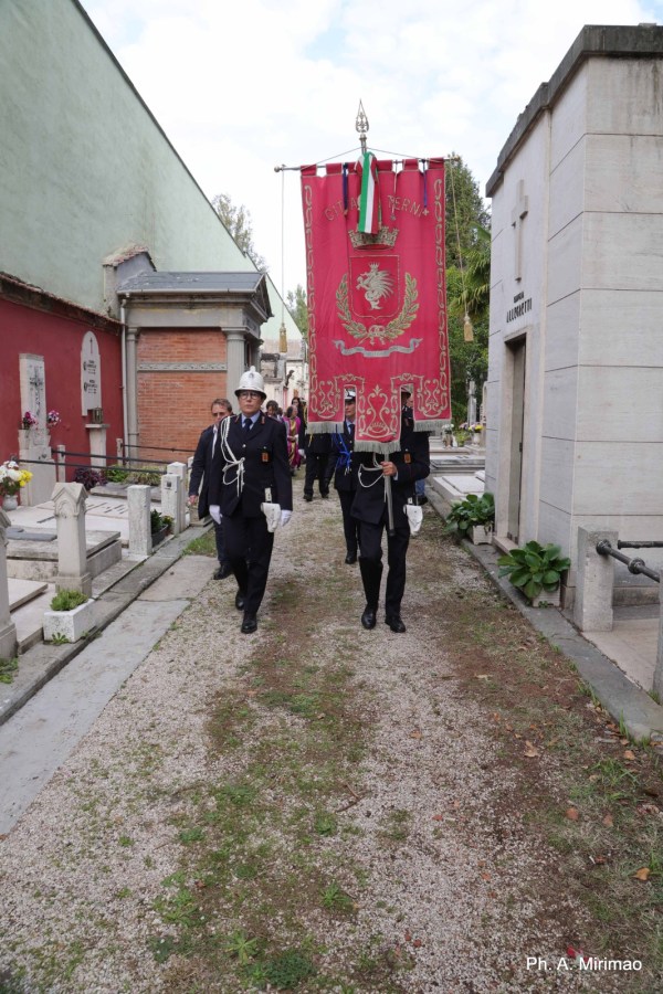 A group of uniformed individuals walking in a cemetery, carrying a large ceremonial banner decorated with a coat of arms and flanked by grave markers.