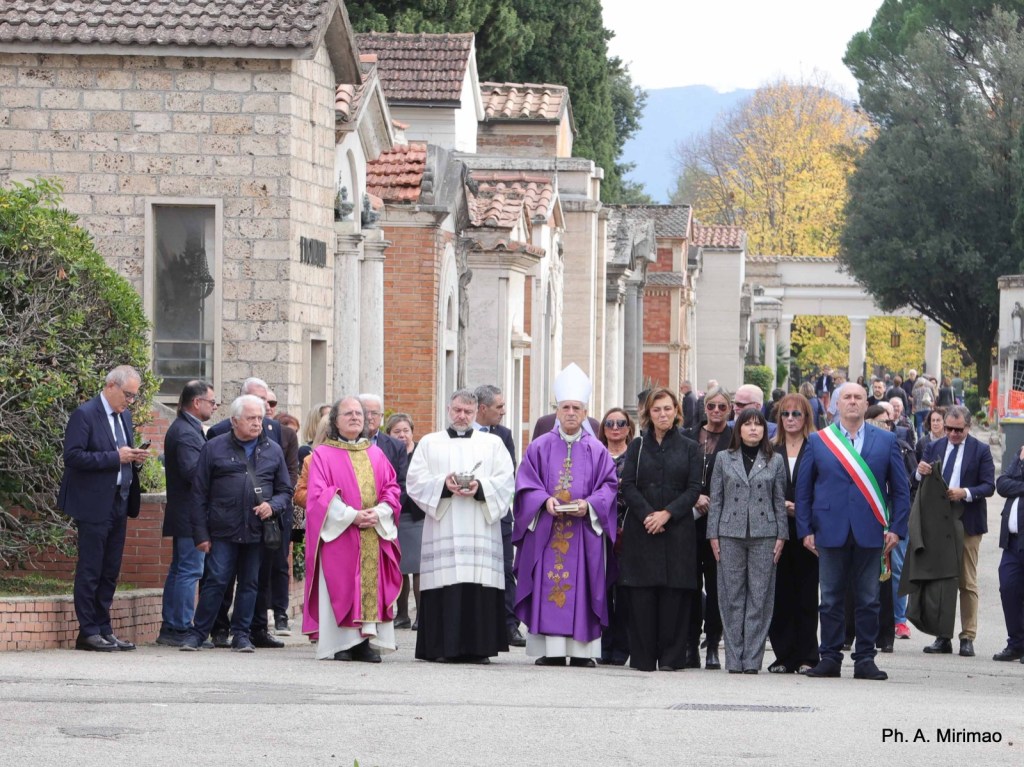 A group of people, including a priest in a purple robe, gathered in a cemetery for a commemorative event. The attendees appear somber and are dressed in formal attire. Several tombs and trees are visible in the background, indicating a respectful and reflective atmosphere.