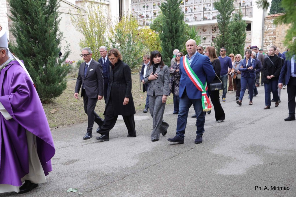 A procession of people walking in a cemetery, with a priest in purple robes leading the group. A man in formal attire wearing a sash walks in the foreground alongside a woman in a gray outfit, while others follow behind.