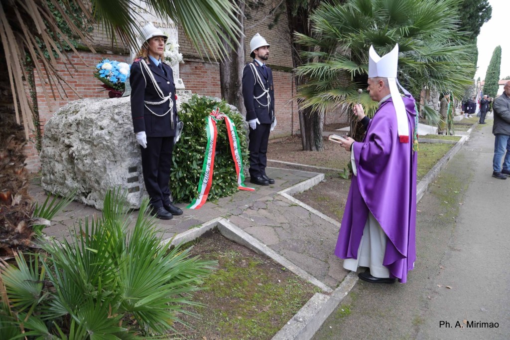 A religious leader in purple robes stands in front of a memorial, while two officers in formal attire stand at attention nearby, surrounded by palm trees and greenery.