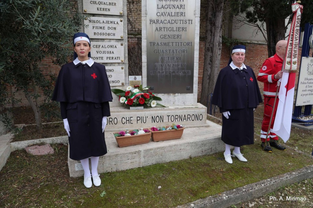 Two women in historical nurse uniforms with red crosses stand at a memorial surrounded by flowers, honoring those who have fallen.