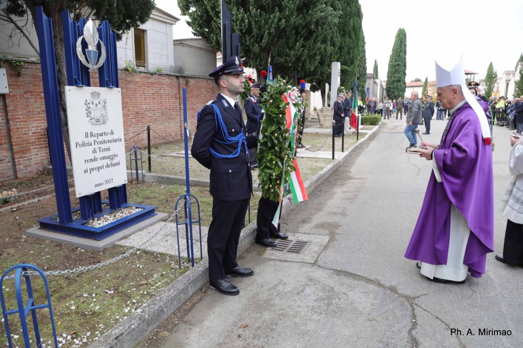 A ceremonial event at a cemetery, featuring a police officer in uniform standing by a memorial sign dedicated to fallen officers. A priest in a purple robe is seen performing a blessing, with a wreath adorned with the Italian flag nearby.