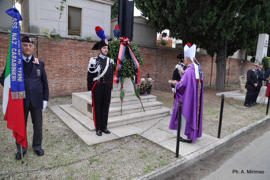 Ceremony honoring fallen soldiers with military personnel, a priest, and wreaths at a memorial site.