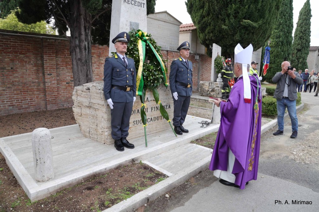 A ceremonial event at a monument with officials in military uniforms and a clergyman in a purple robe, paying tribute with a wreath in a cemetery setting.