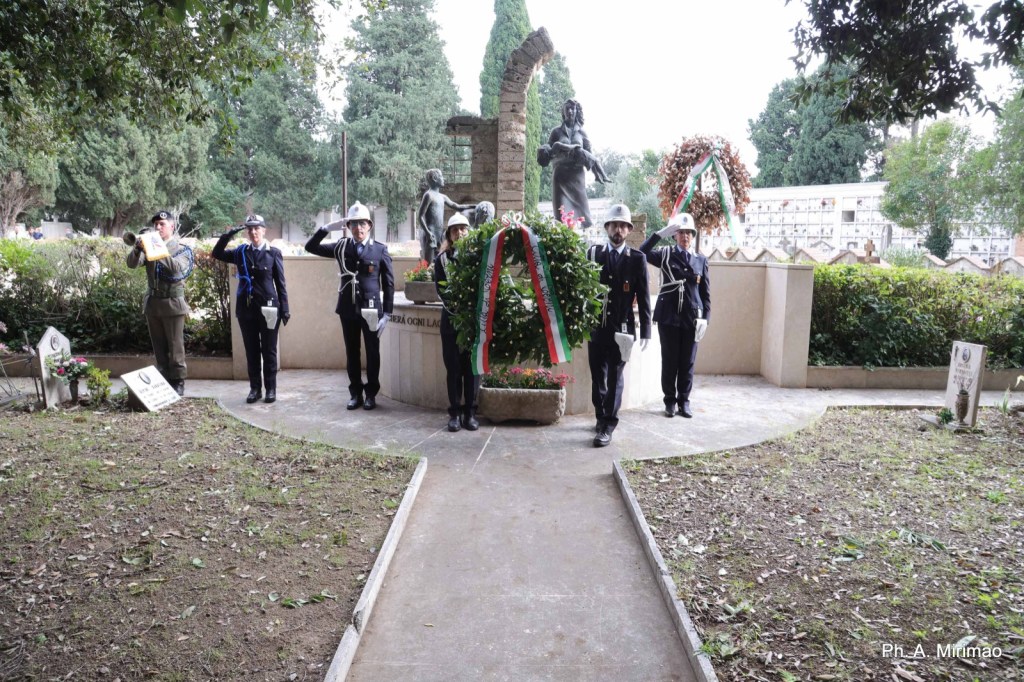 A ceremonial gathering at a memorial site, featuring uniformed personnel saluting and holding a wreath adorned with the Italian flag.