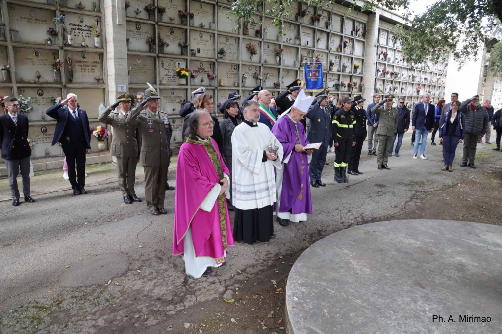 Ceremony at a cemetery with attendees in military attire and clergy, honoring the deceased.
