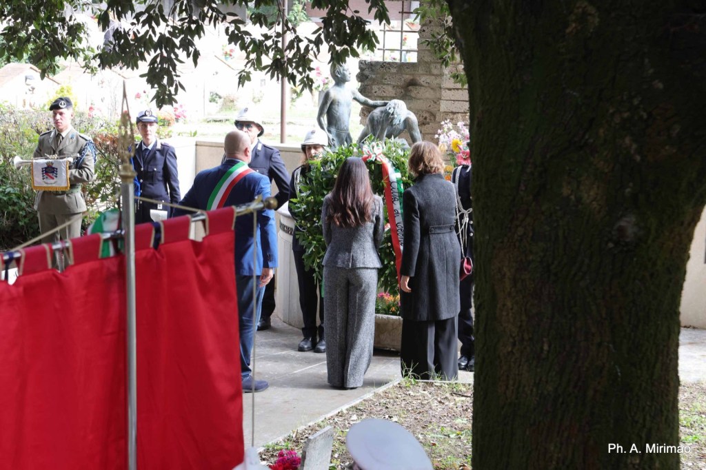 A group of officials and attendees at a memorial ceremony, standing in front of a statue and floral wreaths. Some individuals are in military uniforms while others wear formal attire. Red and green flags are visible in the foreground.