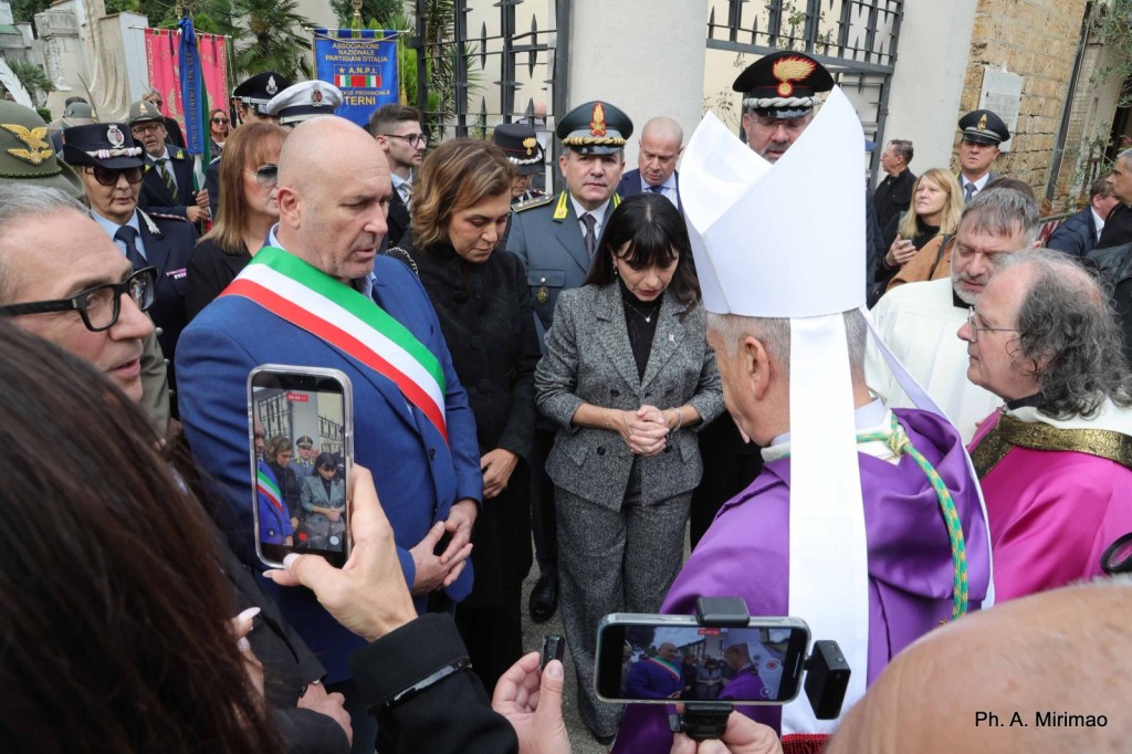 A group of officials and attendees at a formal ceremony, including a man in a suit with a tricolor sash, a church leader in religious attire, and various law enforcement officers, gathered in a respectful moment outdoors.