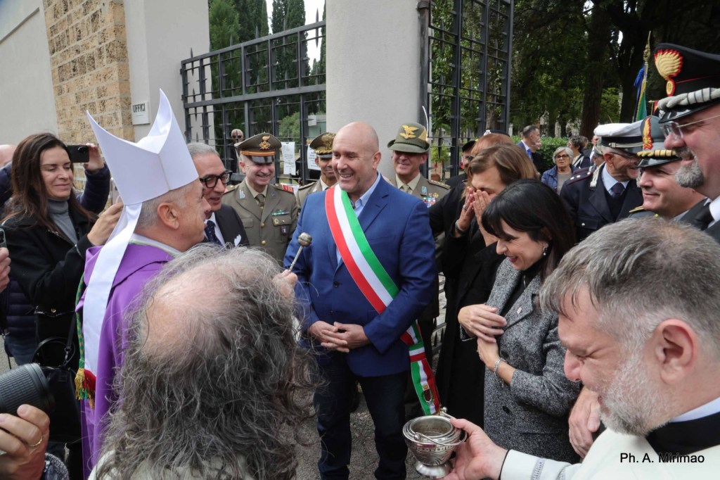A group of people gathered outdoors, including officials in ceremonial attire, interacting and smiling during an event. One man in the center wears a sash representing his position, while a religious figure in a bishop's attire engages with him.