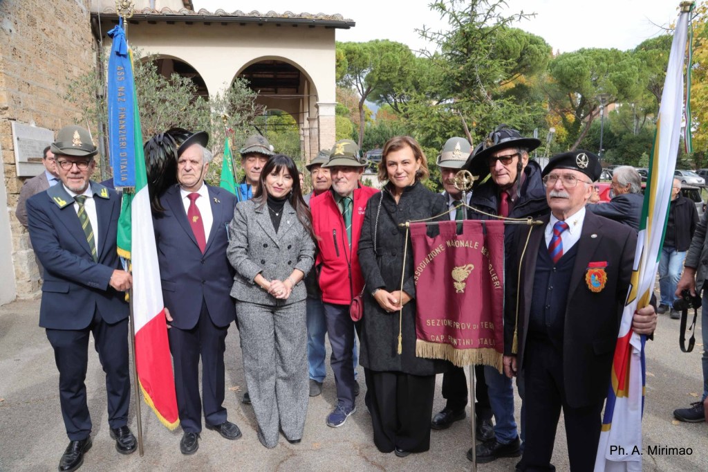 A group of people dressed in formal and traditional attire poses together outdoors, holding flags and banners, set against a backdrop of greenery and architecture.