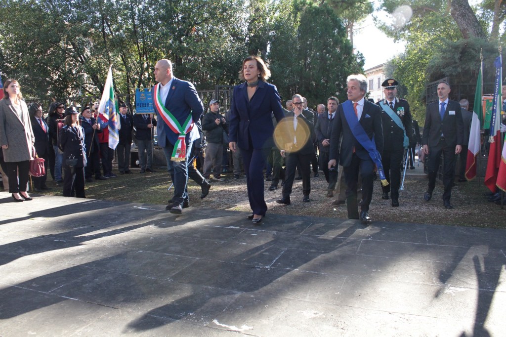 Group of officials walking on a stage during a ceremonial event, surrounded by flags and an audience.