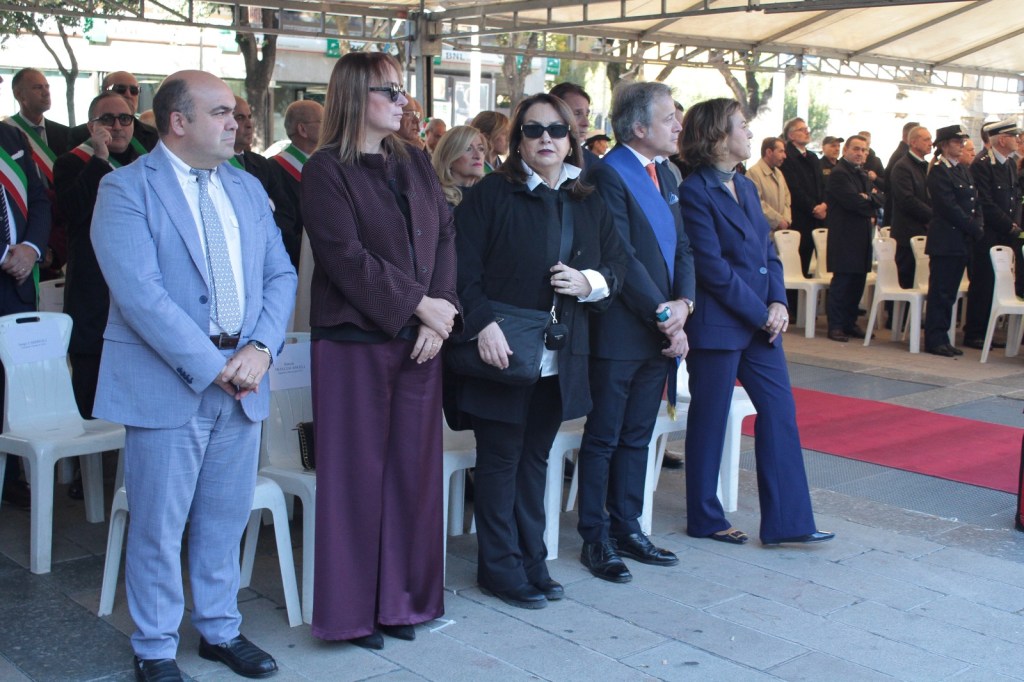 A group of individuals in formal attire standing during a ceremony, with a row of empty chairs in the background.