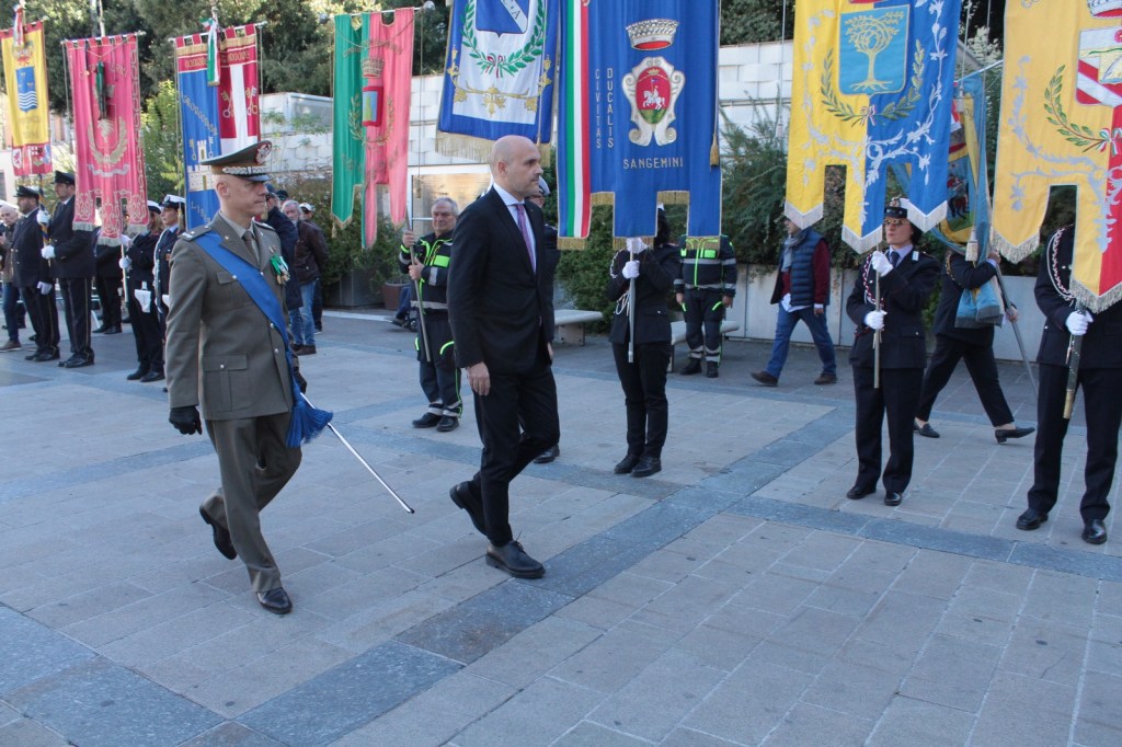 A military officer in uniform and a man in a suit walking during a ceremony, with colorful flags displayed in the background and uniformed personnel standing at attention.