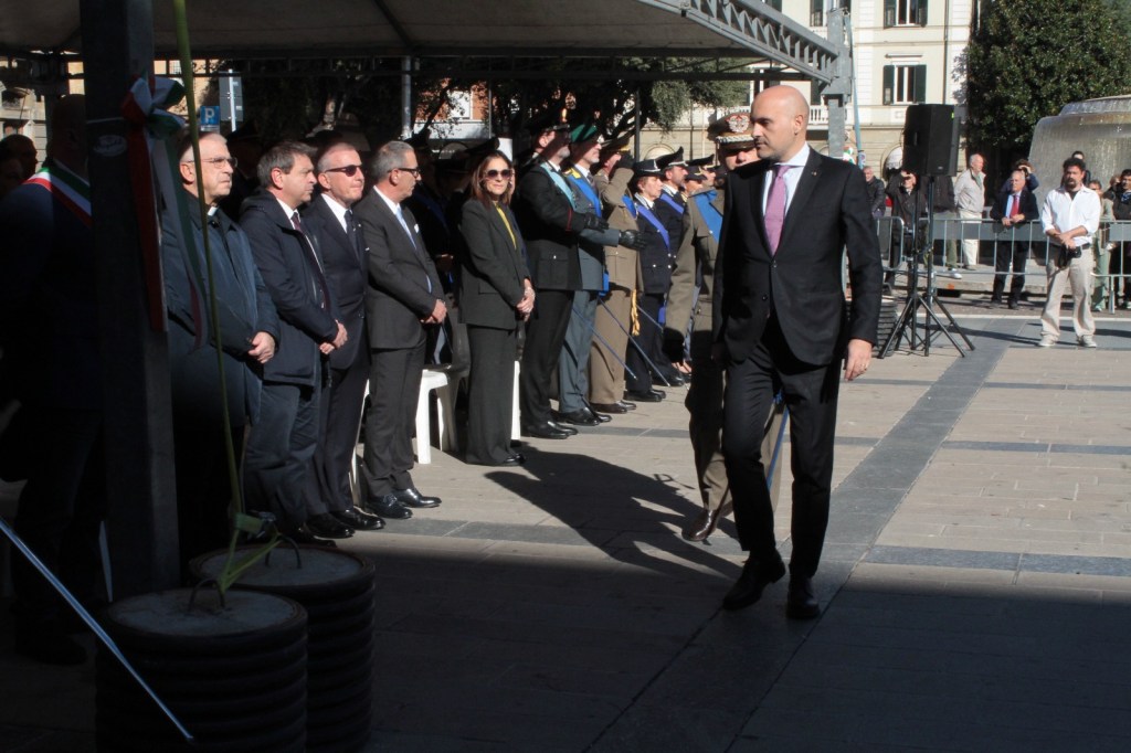 A group of people in formal attire at a commemorative event, with one man walking past the assembled attendees under a canopy.