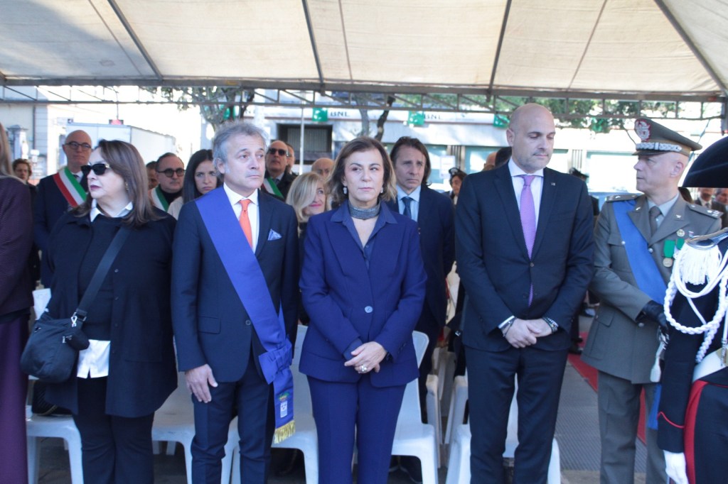A group of formally dressed individuals standing together under a tent, including a man in a blue sash, a woman in a dark suit, and several others in the background, some wearing military uniforms, during a public event.