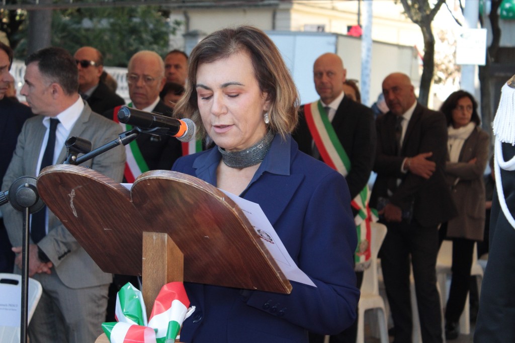 A woman in a blue suit stands at a podium with a microphone, reading from notes. She appears to be at an event attended by several people in formal attire, some wearing tricolor sashes.