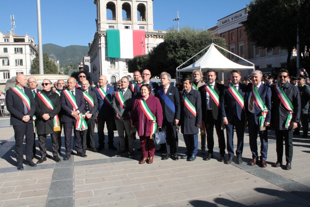 A group of officials wearing sashes in the colors of the Italian flag pose together in a public square, with a large Italian flag displayed in the background.