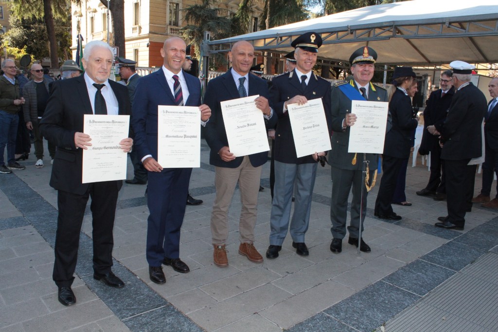 Five men in formal attire holding certificates during a public ceremony outdoors, surrounded by spectators and a tent in the background.