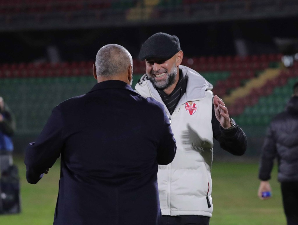 Due uomini sorridenti si abbracciano dopo una partita di calcio, con uno che indossa un cappello e una giacca bianca con il logo della Vis Pesaro, mentre l'altro è in abito scuro.