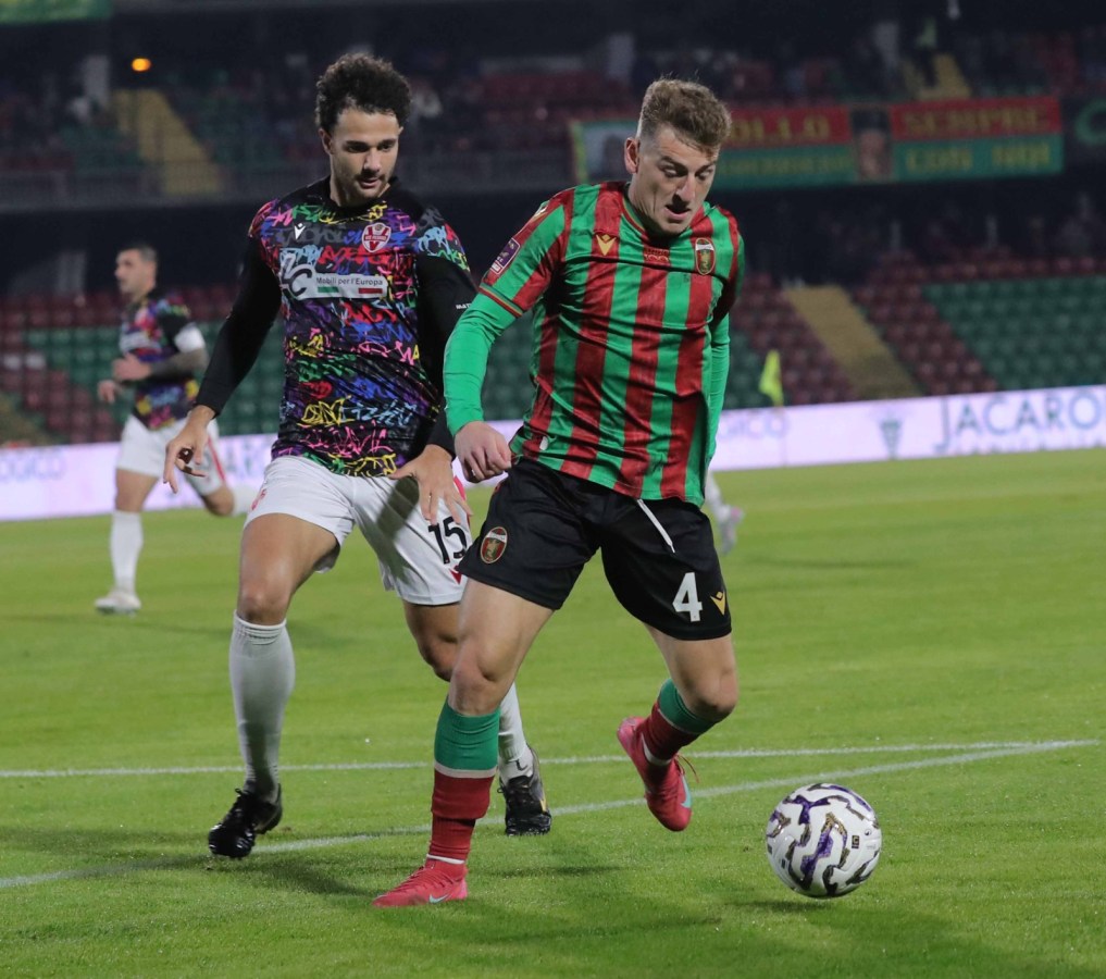 Due calciatori in azione durante una partita di calcio, uno con una maglia a strisce verdi e rosse e l'altro con una maglia colorata e pantaloni bianchi.