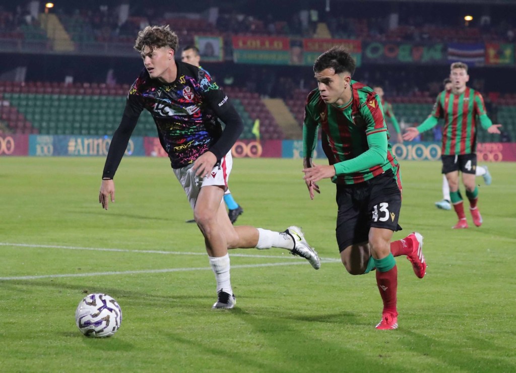 Due calciatori in azione durante una partita di calcio, uno in maglia scura e l'altro in maglia rossa e verde, con una palla vicino a loro.