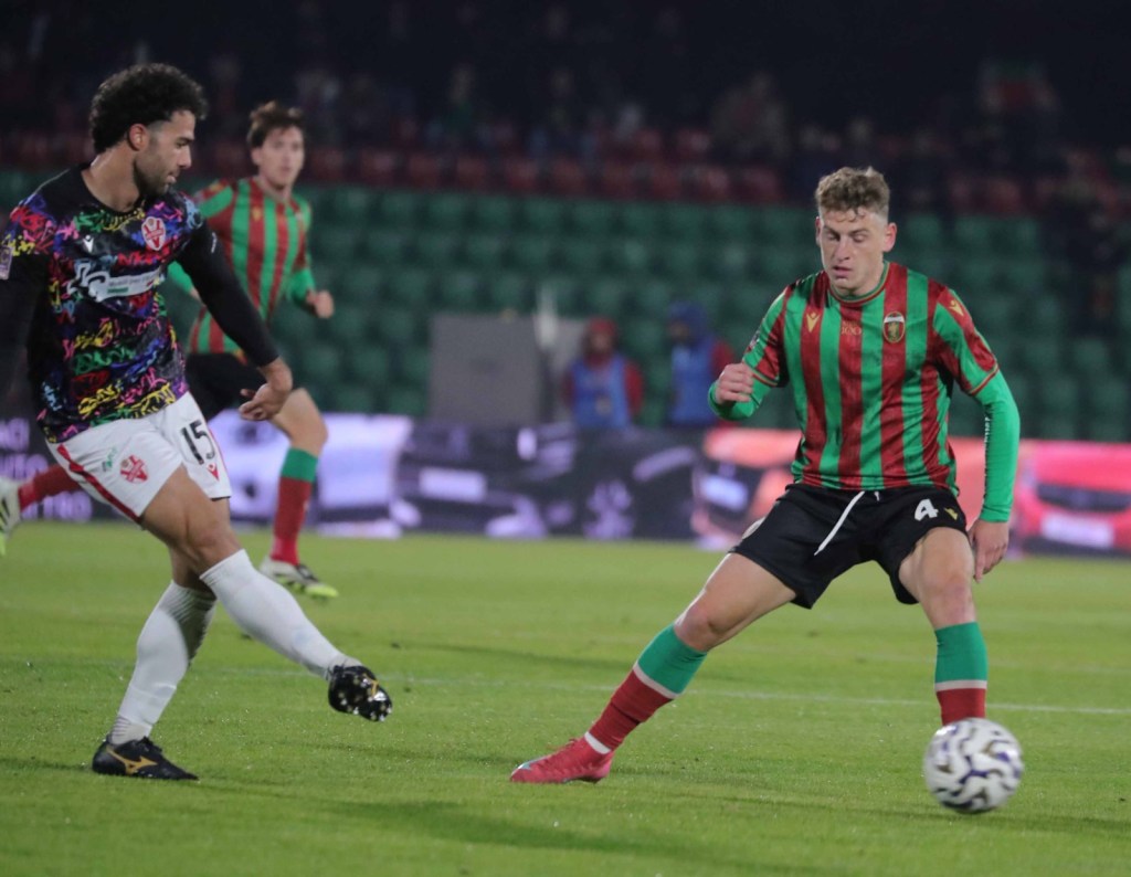 Due giocatori di calcio in azione durante una partita, uno con una maglia colorata e l'altro con una maglia verde e rossa.