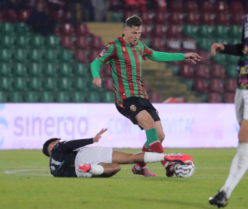 Giocatore della Ternana in maglia rossa e verde intenta a calciare una palla mentre un avversario è a terra sul campo da calcio.