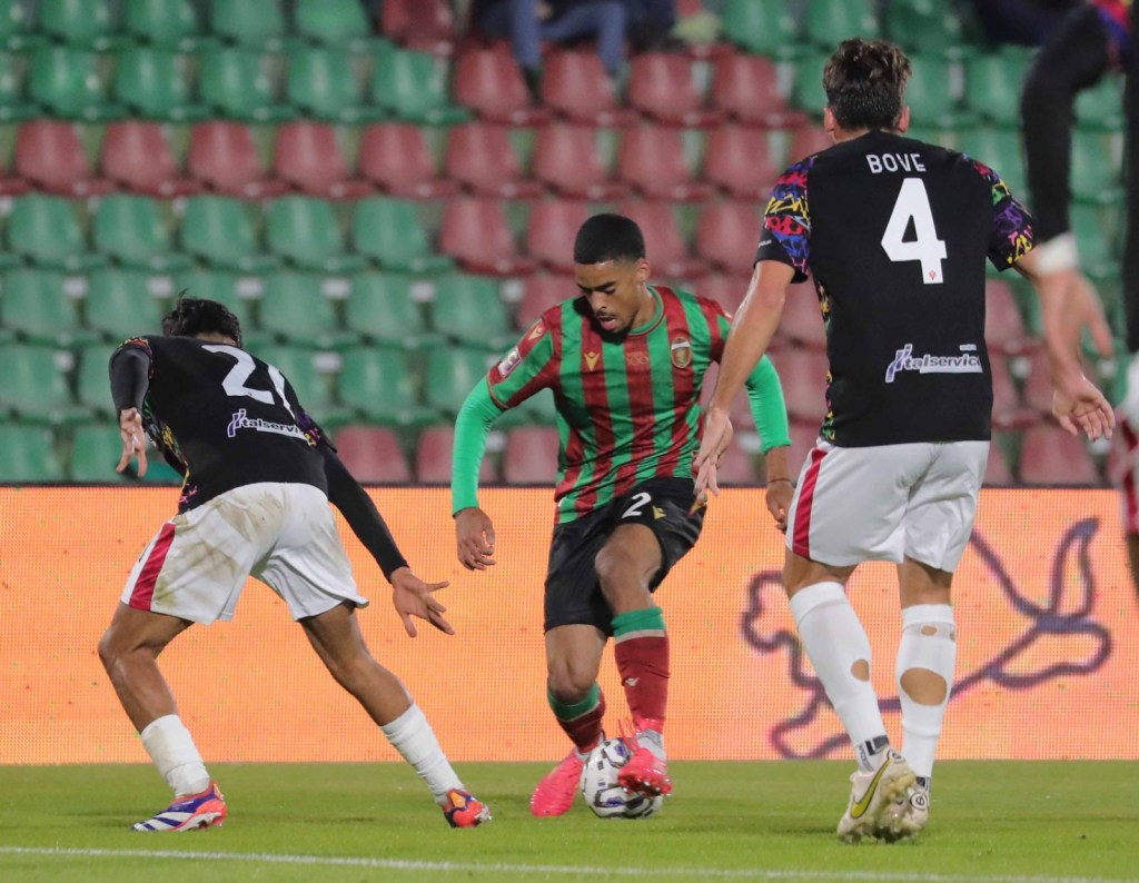 Un momento di gioco in una partita di calcio, con un giocatore in maglia a righe verde e rossa in fase di dribbling, mentre due avversari cercano di contrastarlo.