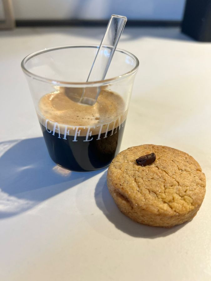 Un bicchiere di caffè espresso con un piccolo biscotto accanto, il tutto posizionato su una superficie chiara.