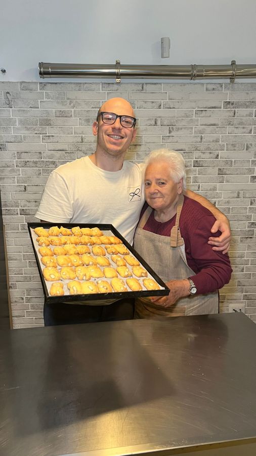 Riccardo Rinaldi e la sua nonna sorridono insieme nella cucina, mentre tengono in mano una teglia con biscotti freschi.
