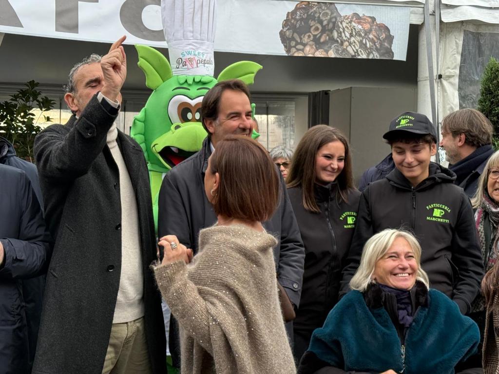 A group of people gathered outdoors, with one man gesturing. A playful green mascot can be seen in the background, wearing a chef's hat. The individuals are wearing winter clothing and appear to be engaged in a lively conversation.