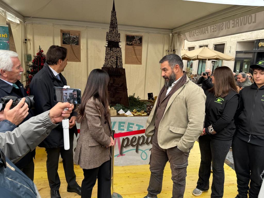 A group of people gathered around a large chocolate sculpture resembling a tower, with a festive atmosphere and decorations in the background.