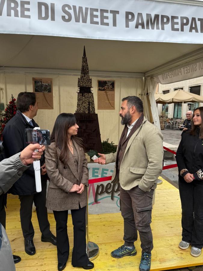 Group of people interacting at a festival booth with a large chocolate sculpture in the background, featuring a decorative sign that reads 'Sweet Pampepatio'.