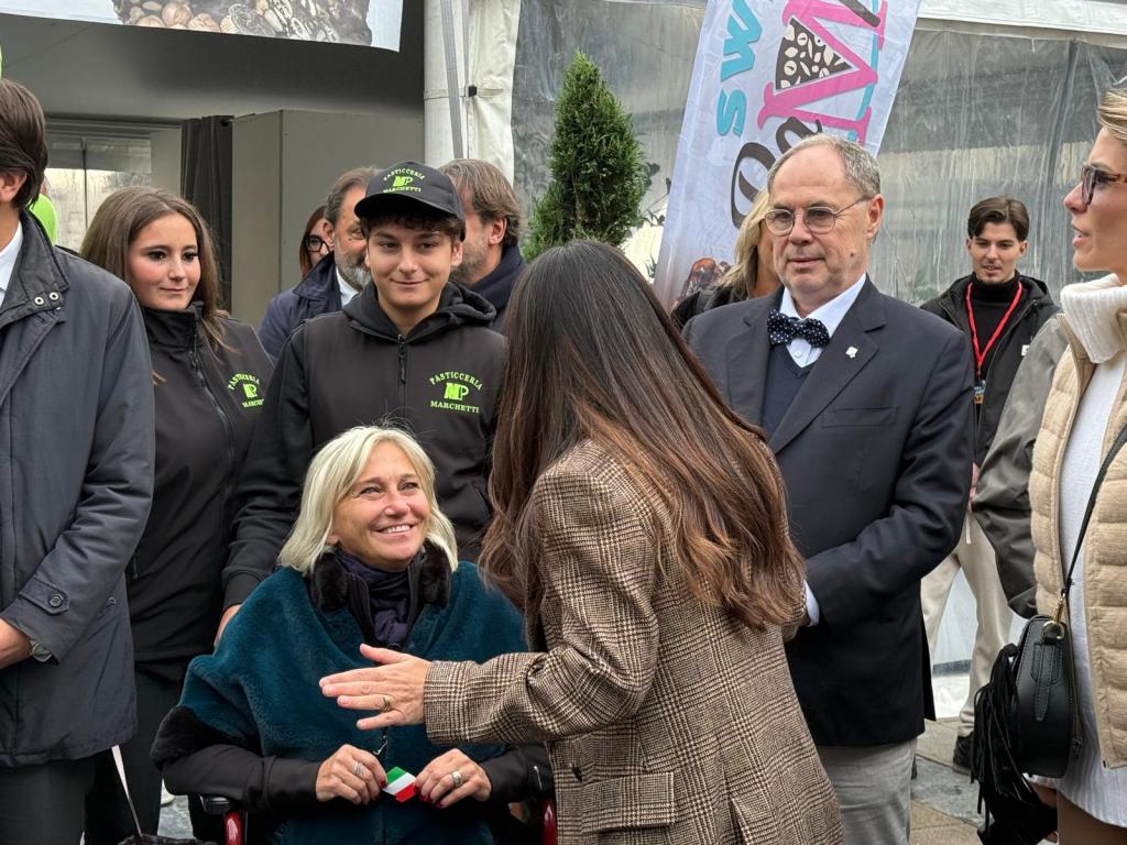 A group of people, including a woman in a wheelchair and several young adults in matching dark jackets, are engaged in conversation in an outdoor setting. The woman seems to be smiling and interacting with another person, while others are observing.