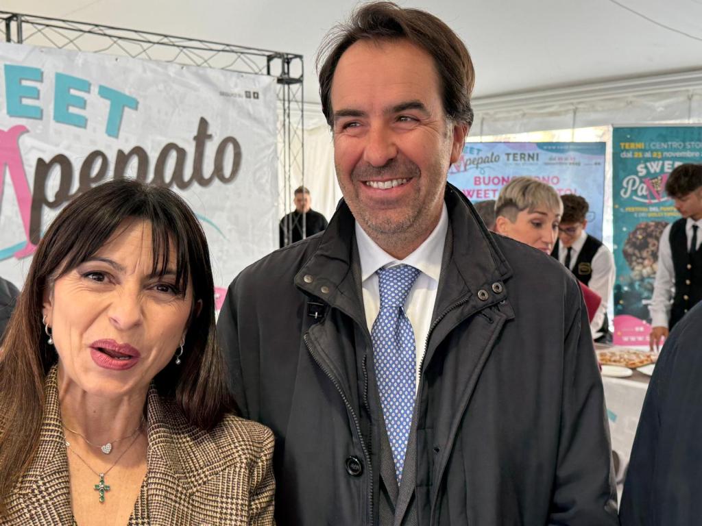 A man and a woman smiling together at an event, with a colorful backdrop displaying the words 'Sweet Pepalto'. The man is wearing a dark coat and a light blue tie, while the woman has long dark hair and a patterned blazer.