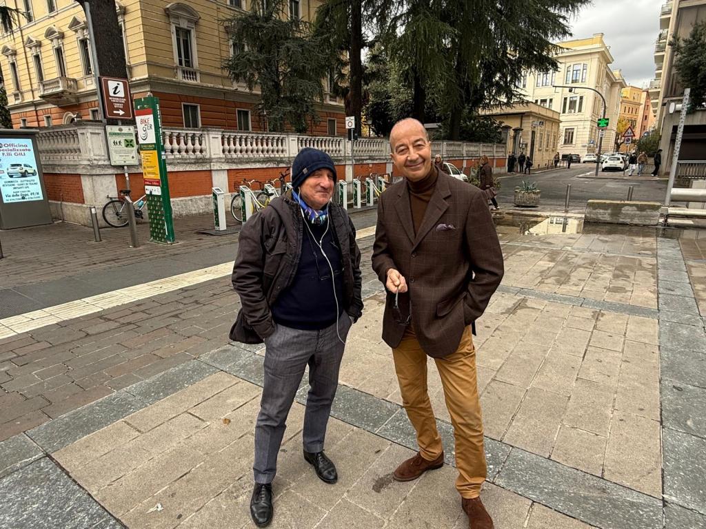 Two men standing on a sidewalk in an urban area, one wearing a dark coat and headphones, the other dressed in a brown suit and holding sunglasses.
