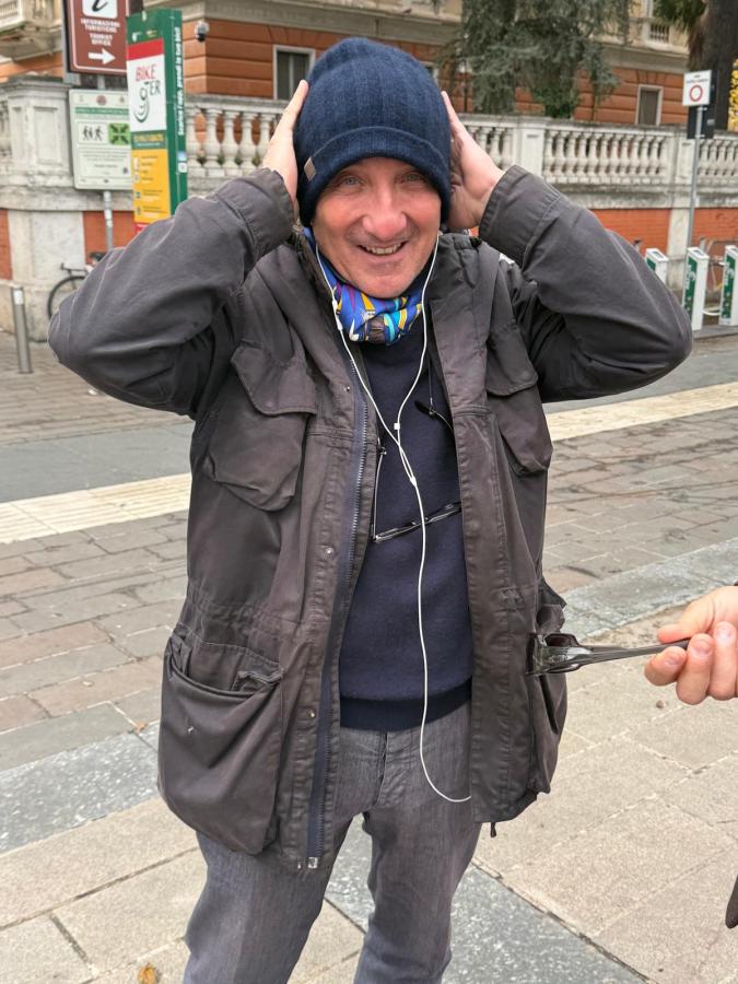 A smiling man with a blue beanie and scarf, holding his hands to his ears, standing outdoors with a city backdrop.