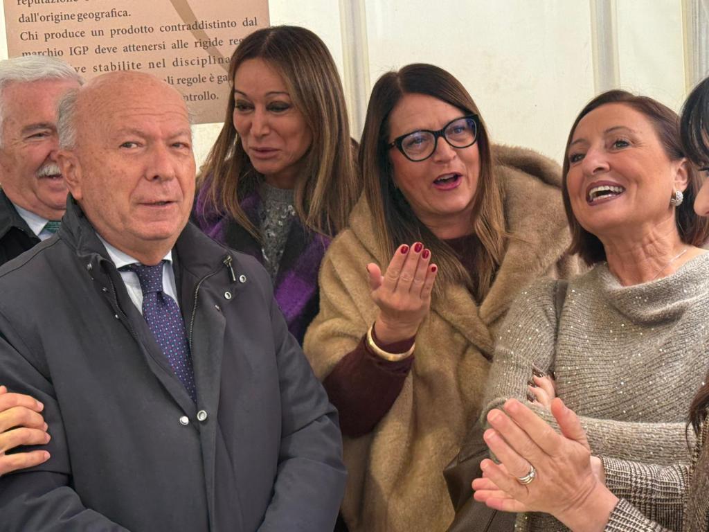 A group of four people, including three women and one man, smiling and engaging in conversation. The background features a sign with text in Italian.