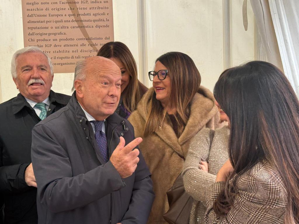 Group of people engaged in conversation, with a focus on a man gesturing while speaking, and several women listening. A sign in the background provides information about a geographic indication.